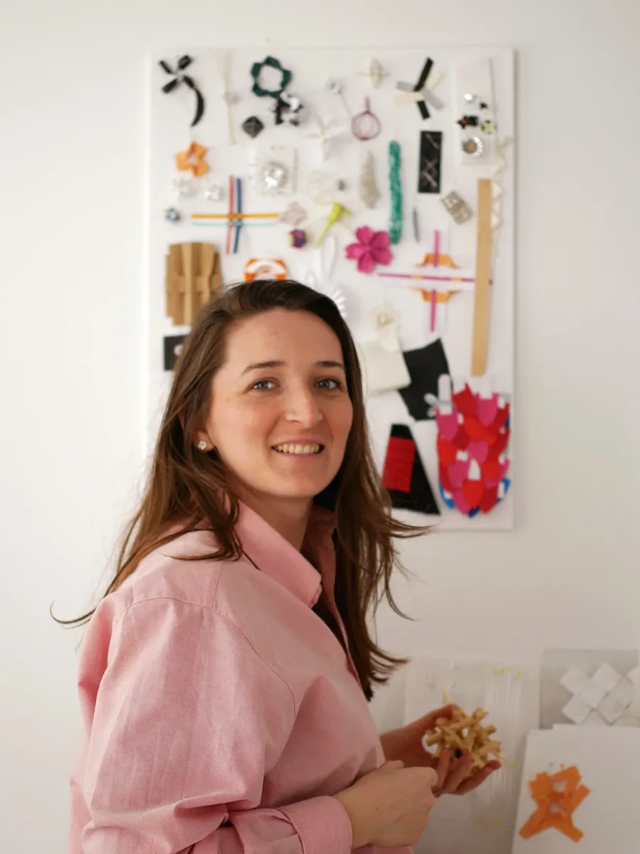 Audrey Bigouin, a talented soft materials designer, stands in front of a wall display showcasing various craft materials and small project samples. She is smiling and holding a handmade craft item in her hands.